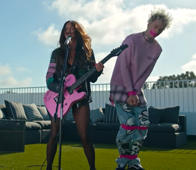 Megan at the microphone, with fishnet tights and holding a pink guitar, and MGK standing next to her with his ankles, knees, and wrists tied up with pink tape