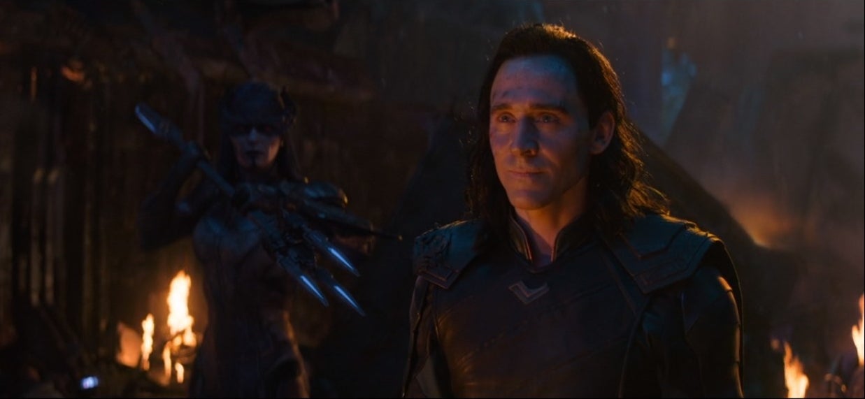 Loki stands is being guarded by a woman near a fire