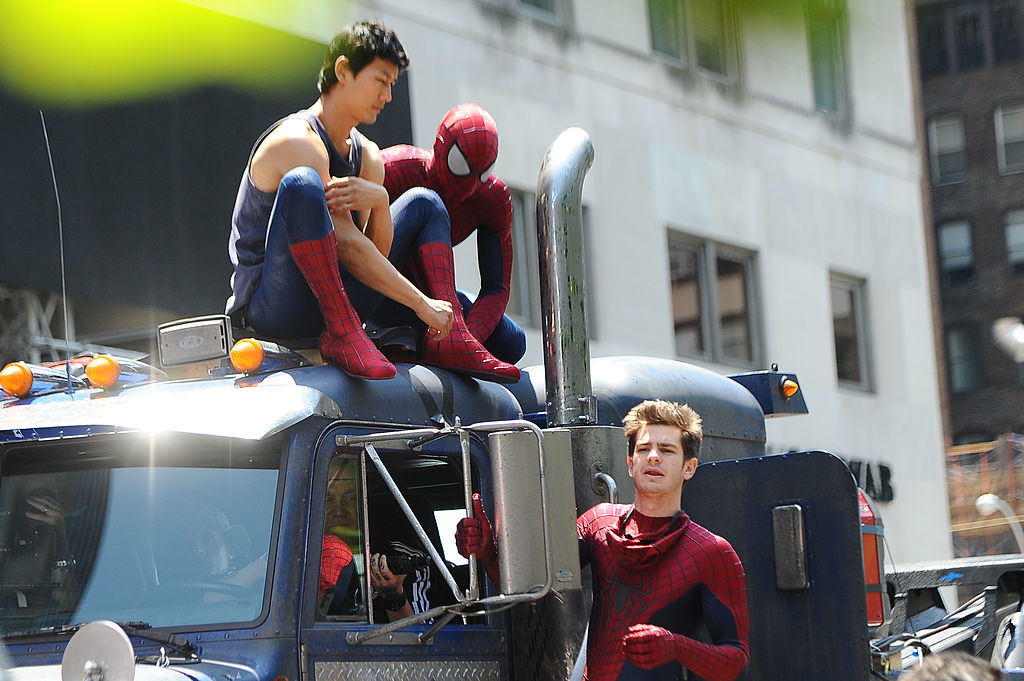 Andrew Garfield on the side of truck while two other men dressed in the same Spider-Man costume stare down at him from their seated position on top