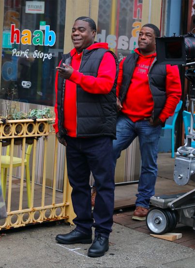 Tracy Morgan standing on set in a red hoodie and black vest; a man who looks like him is standing behind him wearing the same
