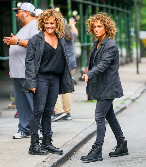 Jennifer Lopez dressed in work boots, skin tight, dark jeans, and a dark jacket standing next to a double in her same outfit