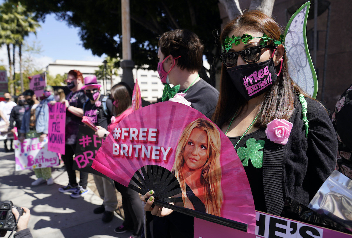 A person wears a face mask and holds up a handheld fan, both of which read &quot;#FreeBritney&quot;