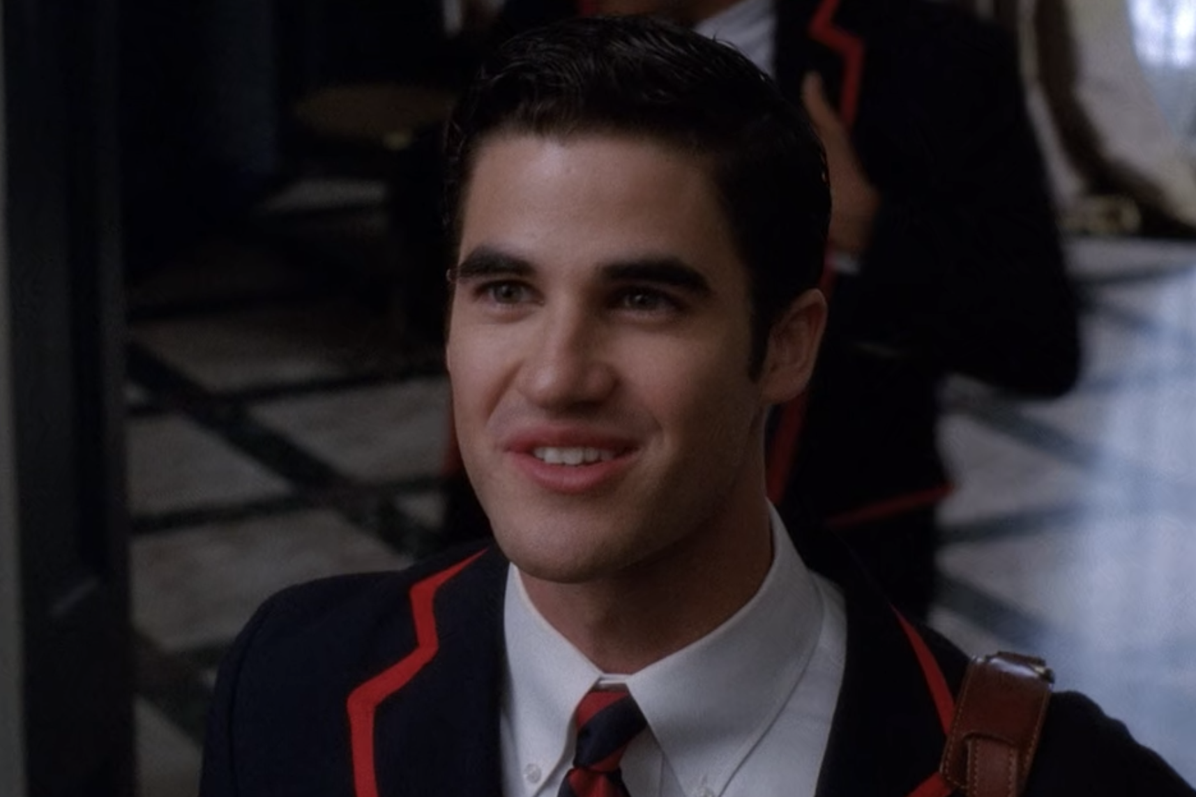 A close up of Blaine Anderson as he wears his blue and red school uniform blazer and smiles up at Kurt Hummel.