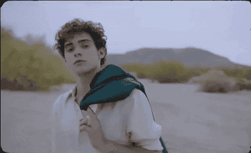 Joshua Bassett, holding a sweater over his shouder, looks brooding in the desert in this gif