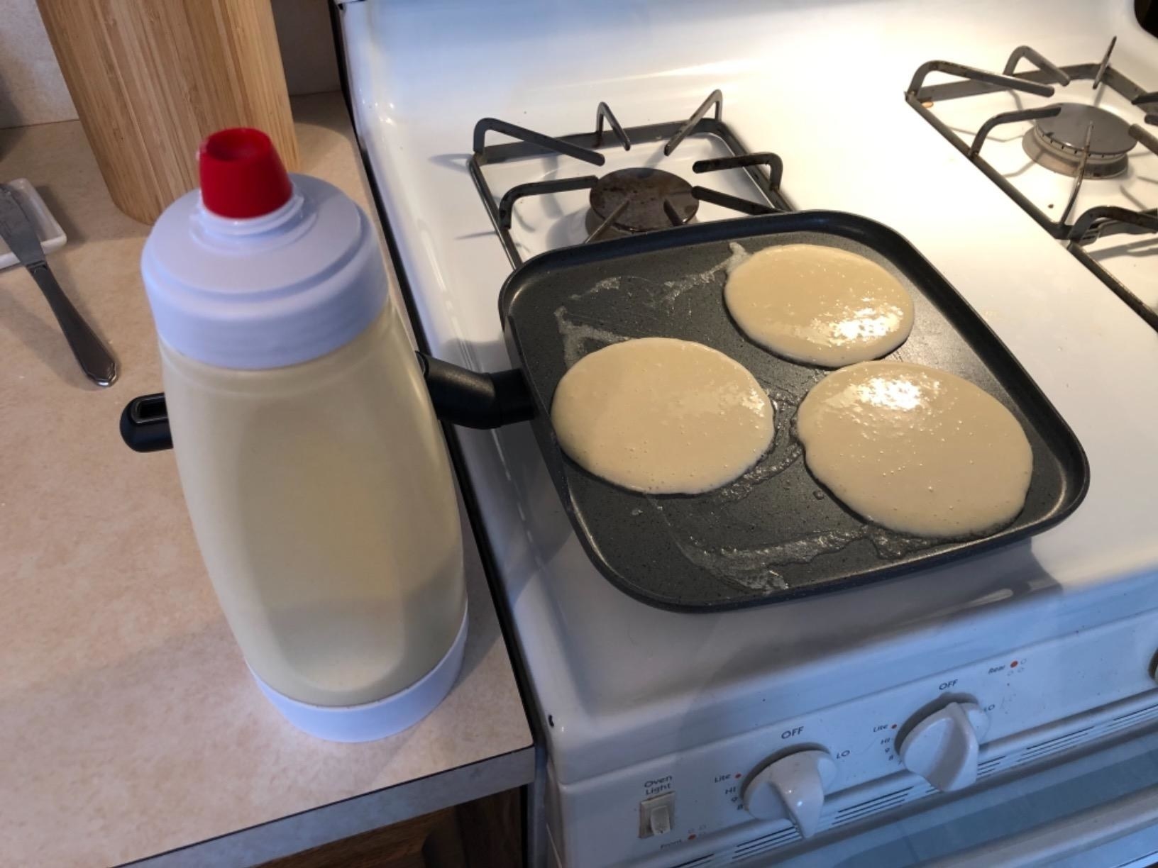 the pancake batter mixer and dispenser with batter inside next to a skillet with freshly poured batter cooking into pancakes on the stove