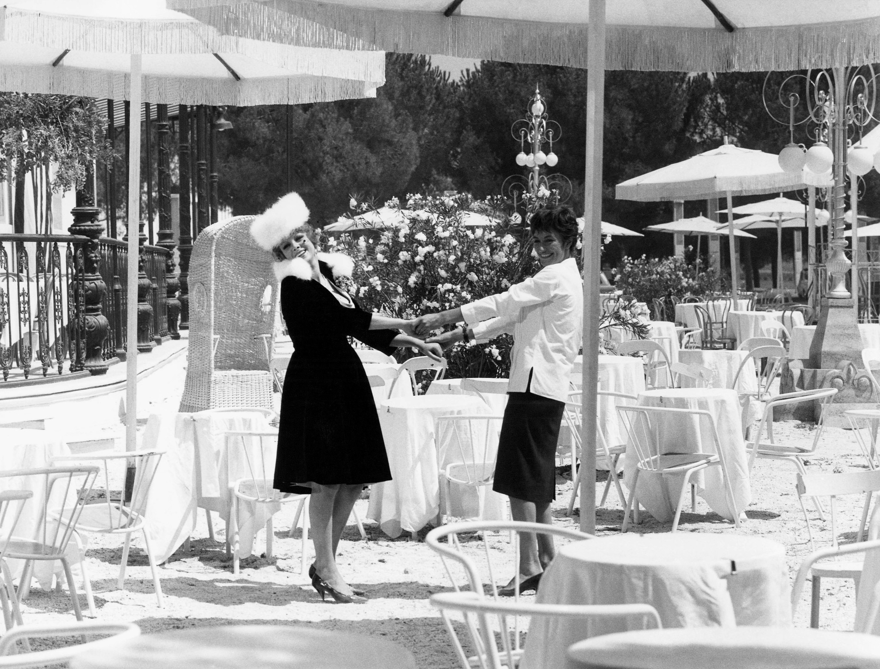 Black and white: Two women in stylish outfits standing with arms stretched toward one another, holding hands at a beautiful outdoor dining area with tables, chairs, and fringed umbrellas, still from 8 1/2