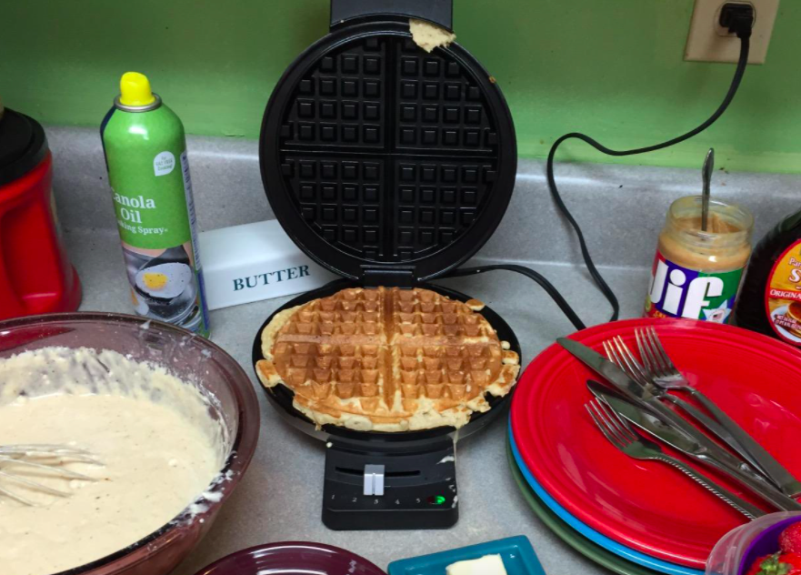 A customer review photo of the waffle maker filled with a cooked waffle on their kitchen counter surrounded with the tools to make and serve waffles