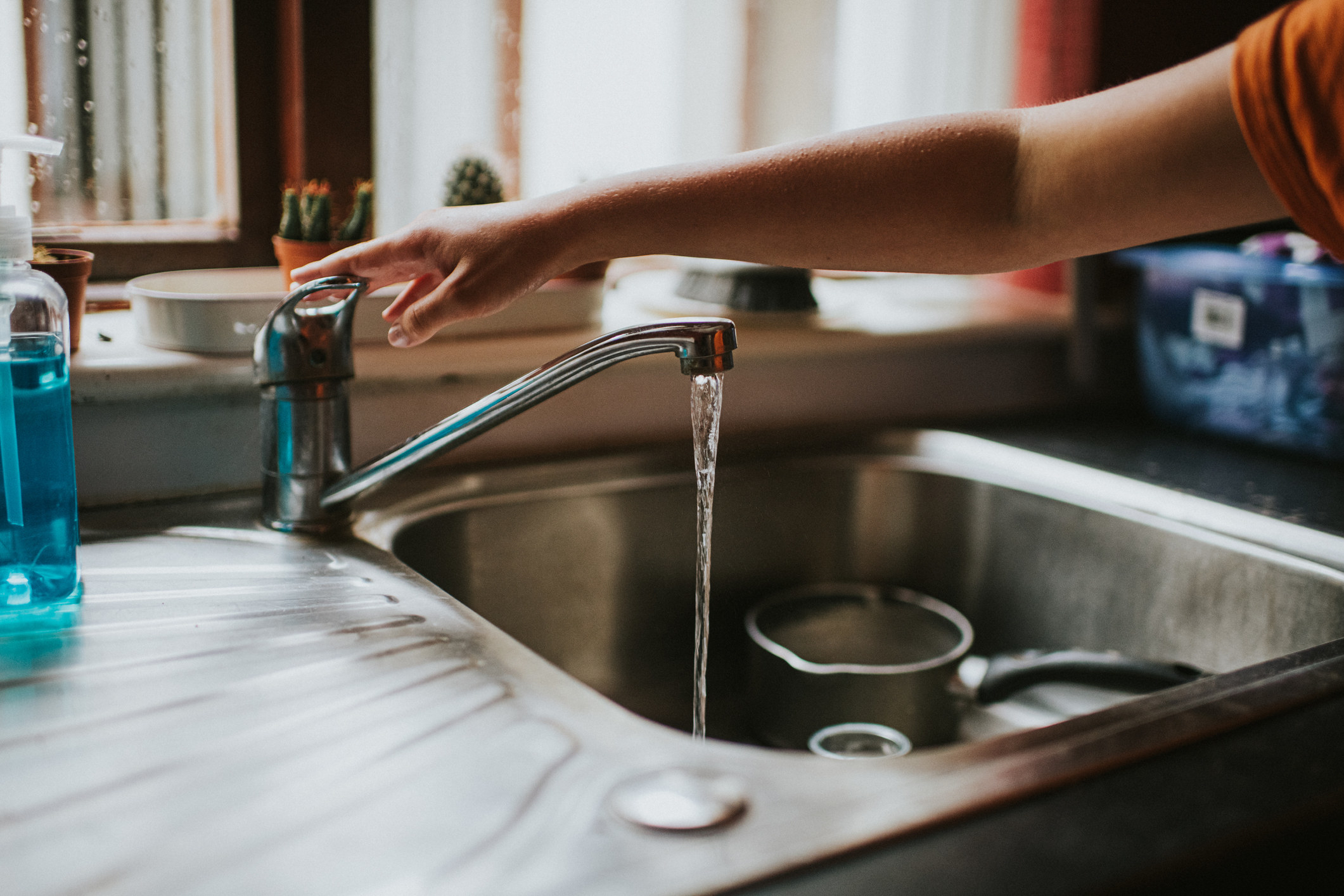 An arm pushing down the handle of a running faucet with a small pot and a glass in the sink