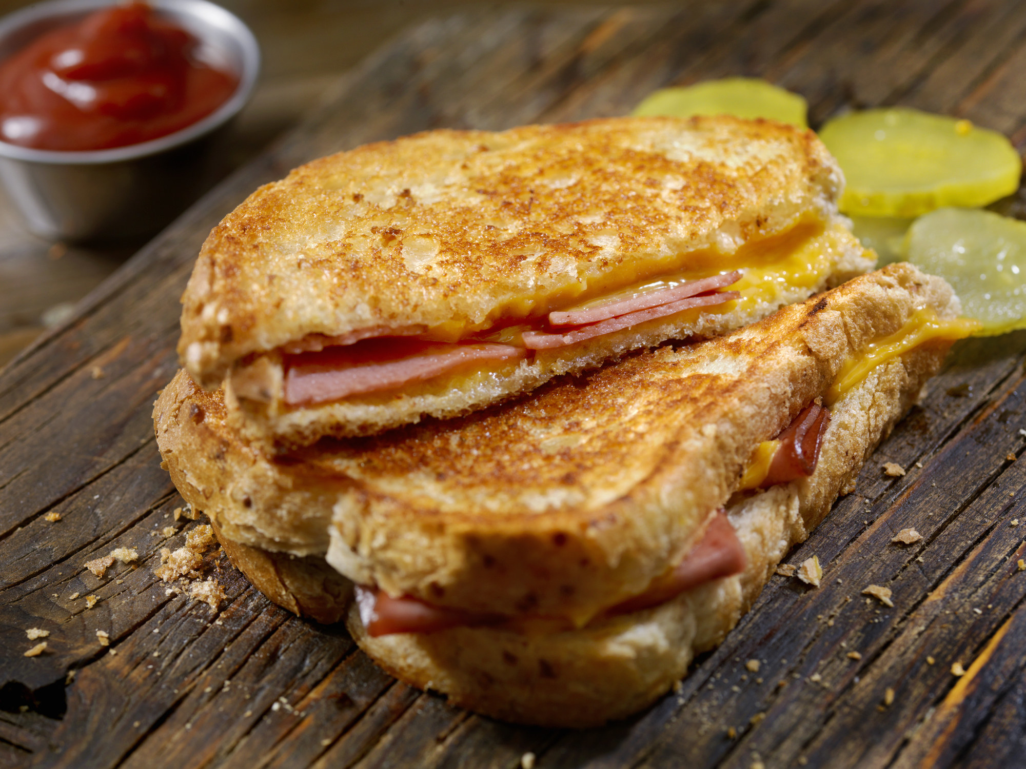 Grilled cheese and bologna sandwich on a wooden board with pickle slices and a metal ramekin of ketchup in the background