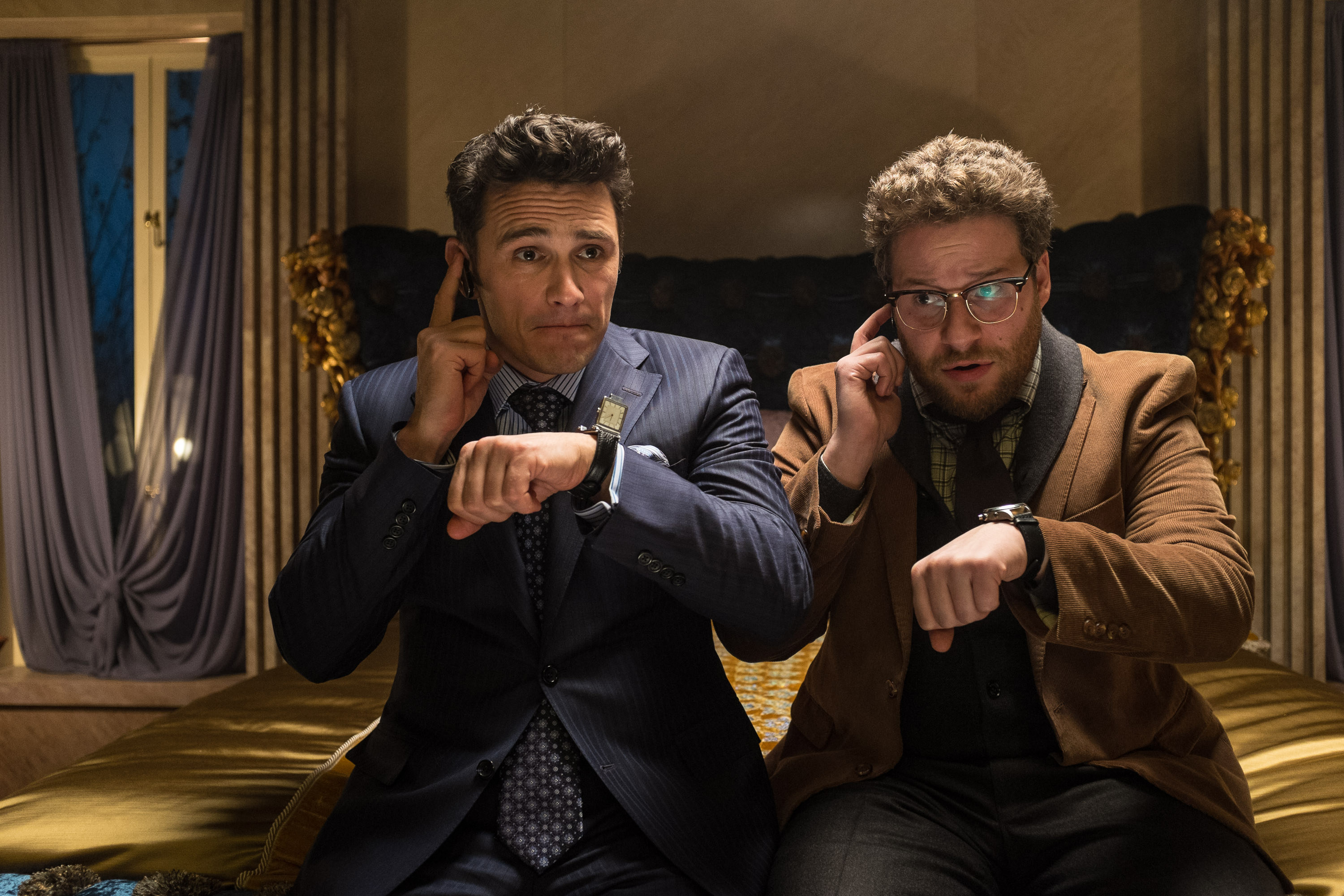 James Franco and Seth Rogen sit next to each other while touching their respective earpieces