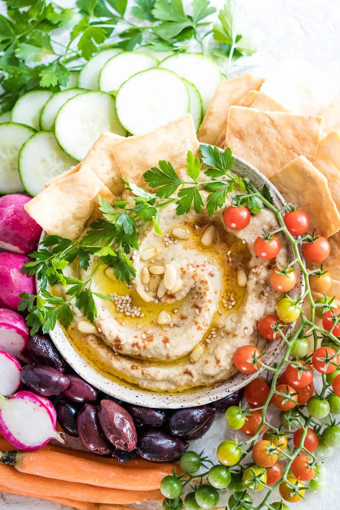 A creamy dip topped with oil, nuts, seeds, and herbs surrounded by colorful veggies and pita chips