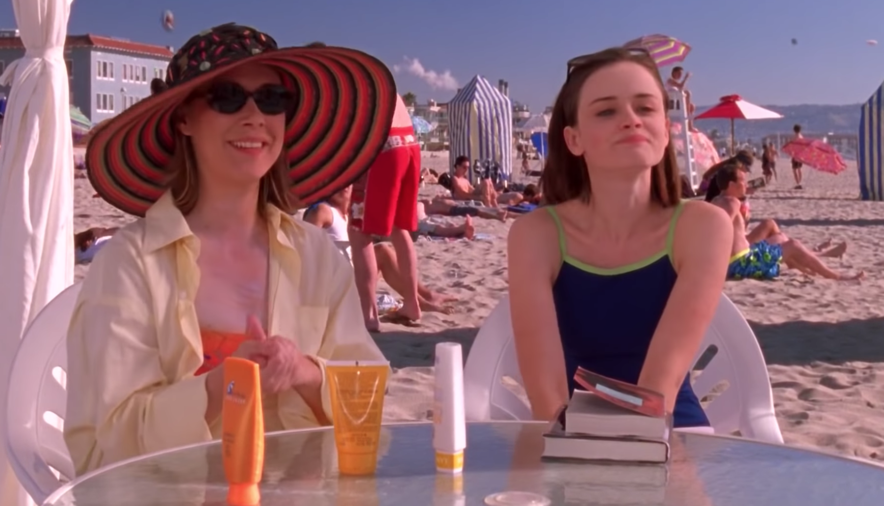 Paris Geller wears an unbuttoned yellow shirt and black-and-orange striped sun hat, and Rory Gilmore wears a blue one-piece swimsuit with green straps