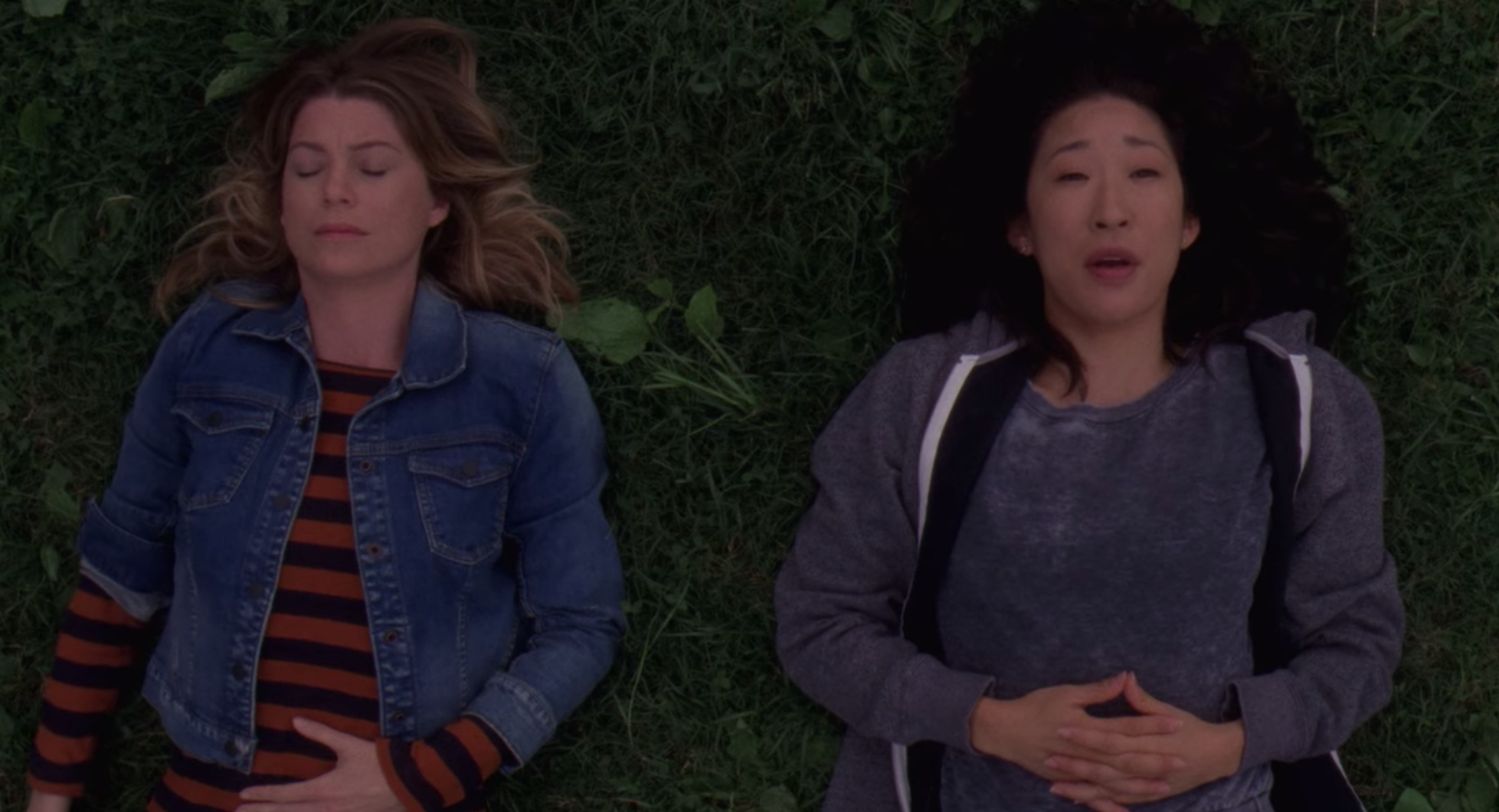Meredith Grey, with her eyes closed and wearing a red-and-black striped shirt under a denim jacket, lies on the grass next to Cristina Yang, wearing a gray shirt under a matching sweater