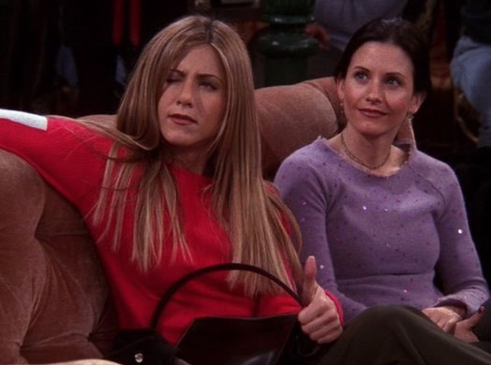 Rachel Green sits on a café couch with an arm thrown over the back with one eye closed as Monica Geller sits beside her.