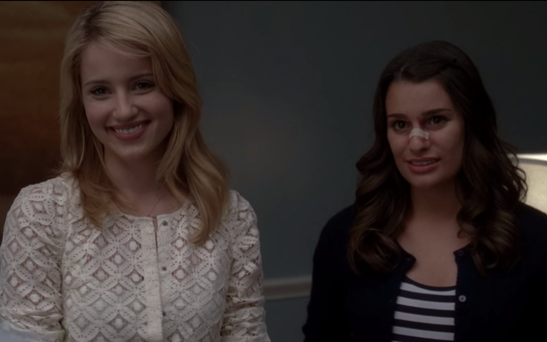 Quinn Fabray, wearing a white lace sweater, smiles brightly while Rachel Berry grimaces as her nose is broken