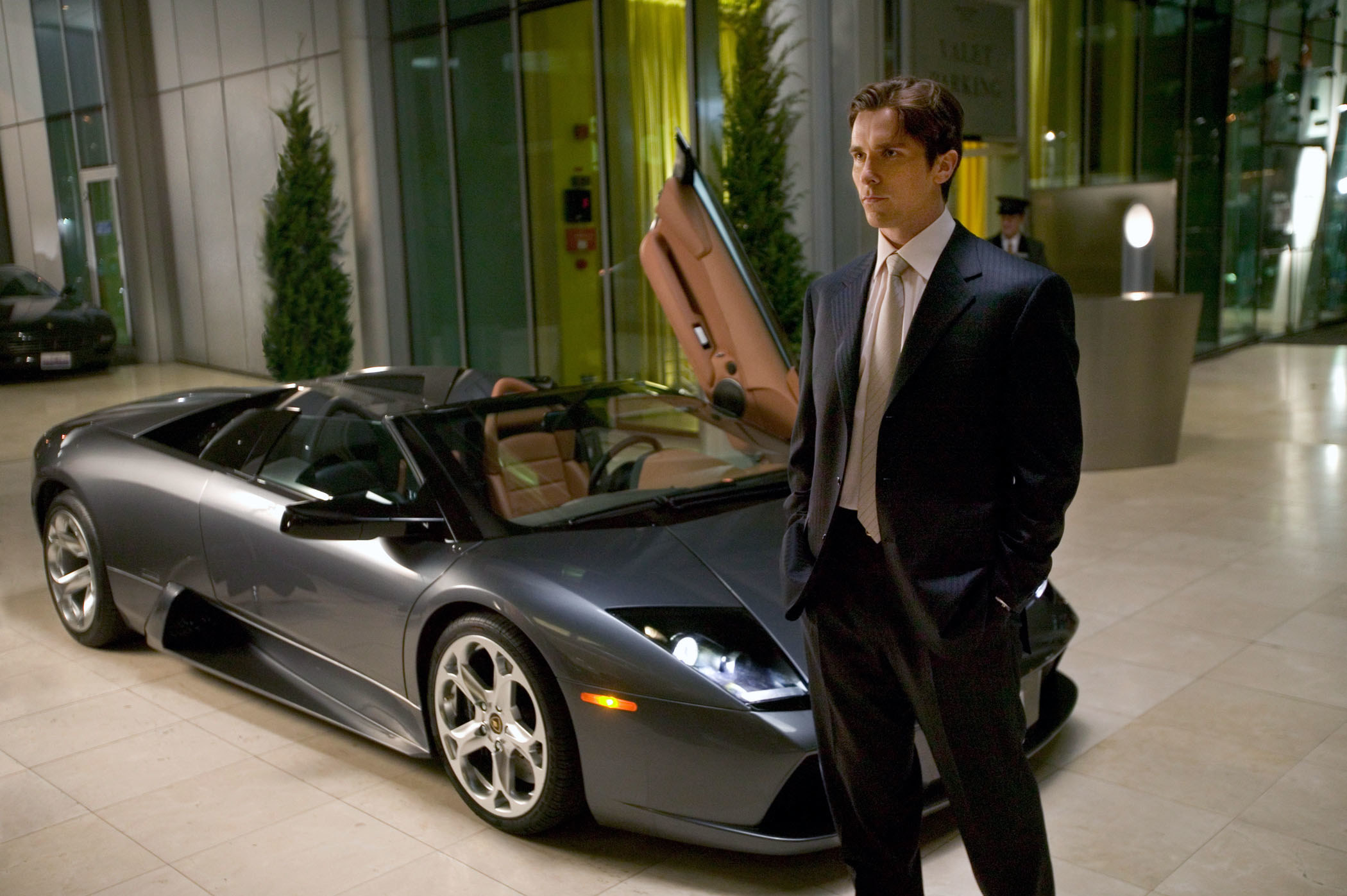Christian Bale as Bruce Wayne standing in front of a fancy car in &quot;Batman Begins&quot;