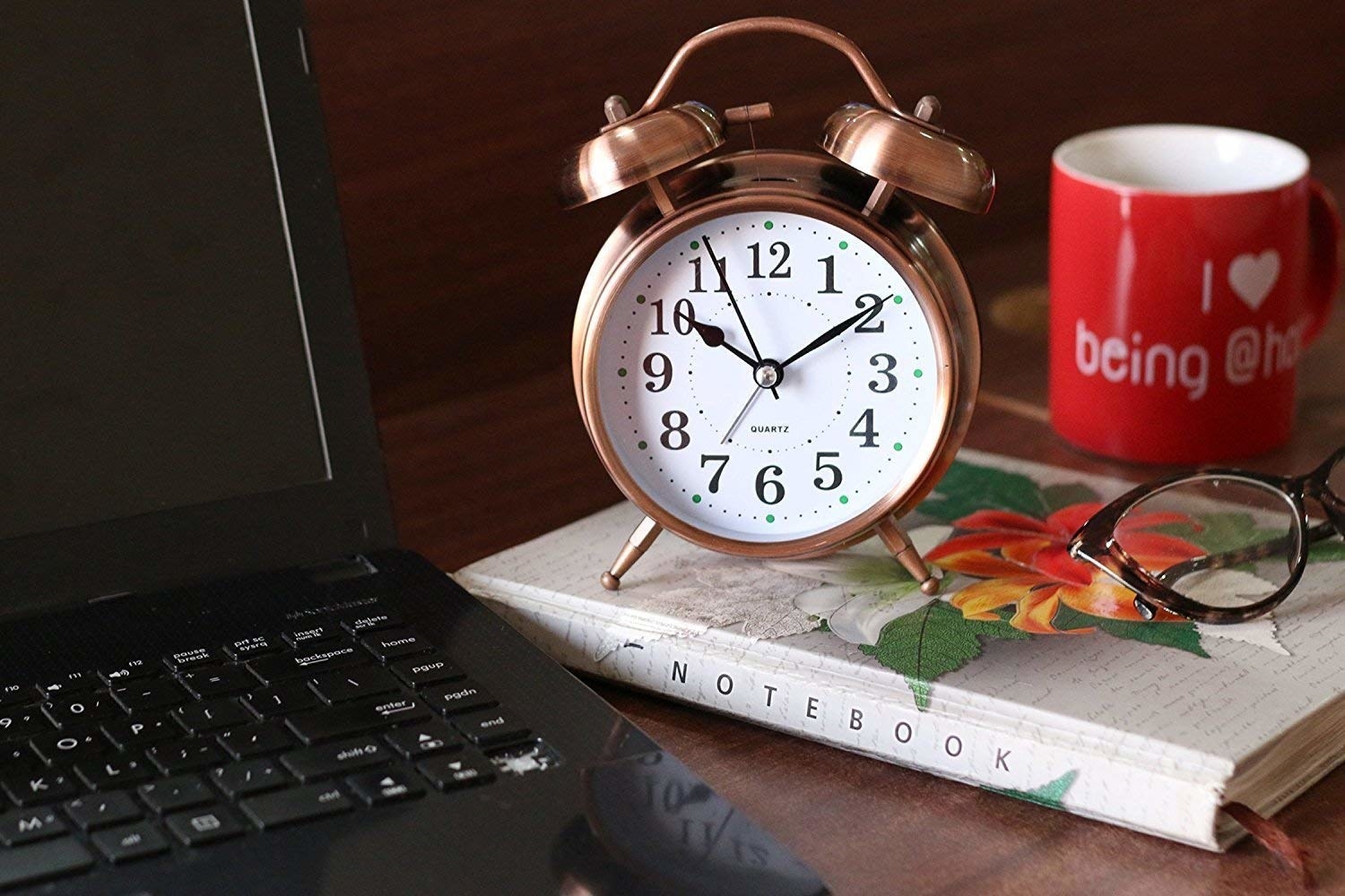 A copper-coloured metal analogue alarm clock kept on a notebook next to a laptop.