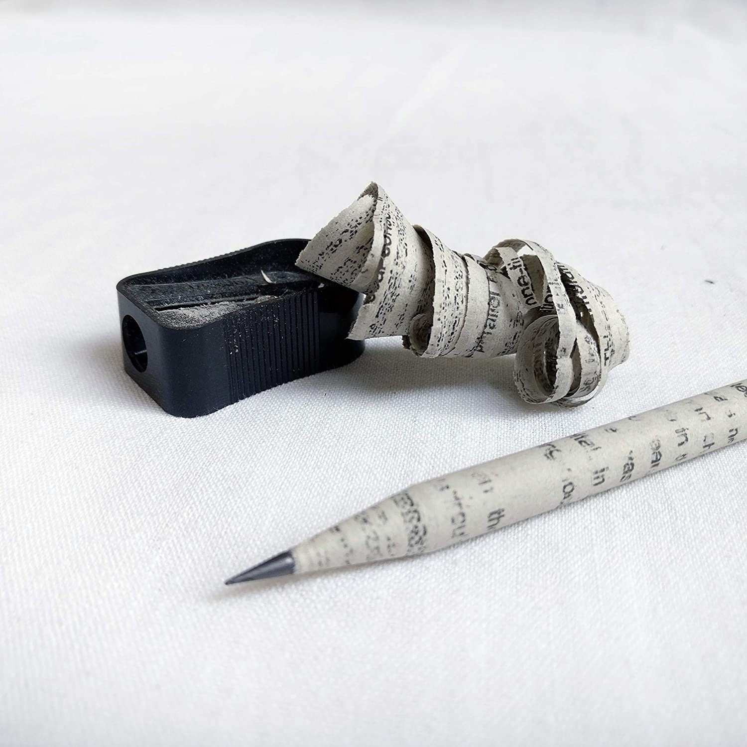 A recycled newspaper pencil pictured with a black sharpener and pencil shavings.
