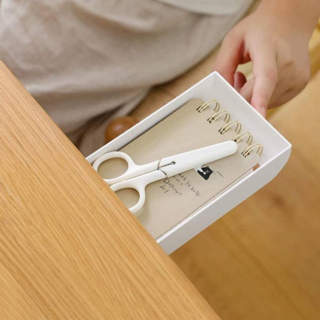 A small drawer pulled open from a desk with a notebook and pair of scissors in it