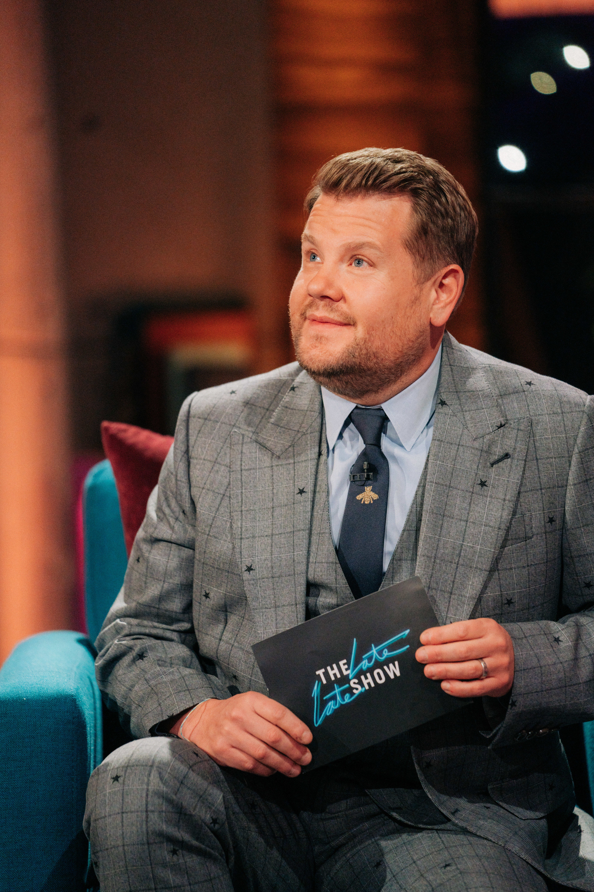 Corden sitting in a chair smiling and holding a &quot;The Late Late Show&quot; card