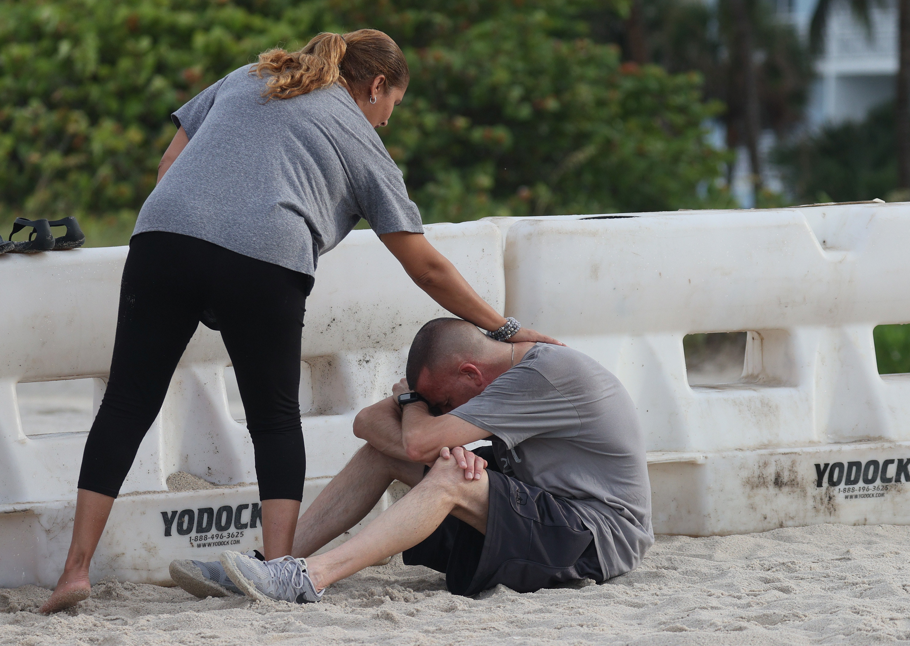 A man sits in the sand with his head in his hands as a woman pats his back