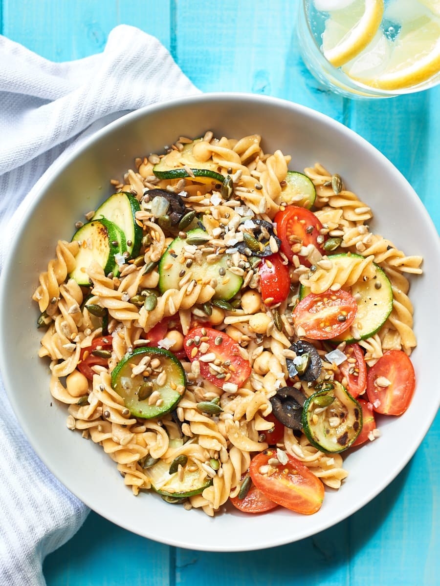 Pasta, tomatoes, zucchini, olives, and nuts in a white bowl on a blue surface.