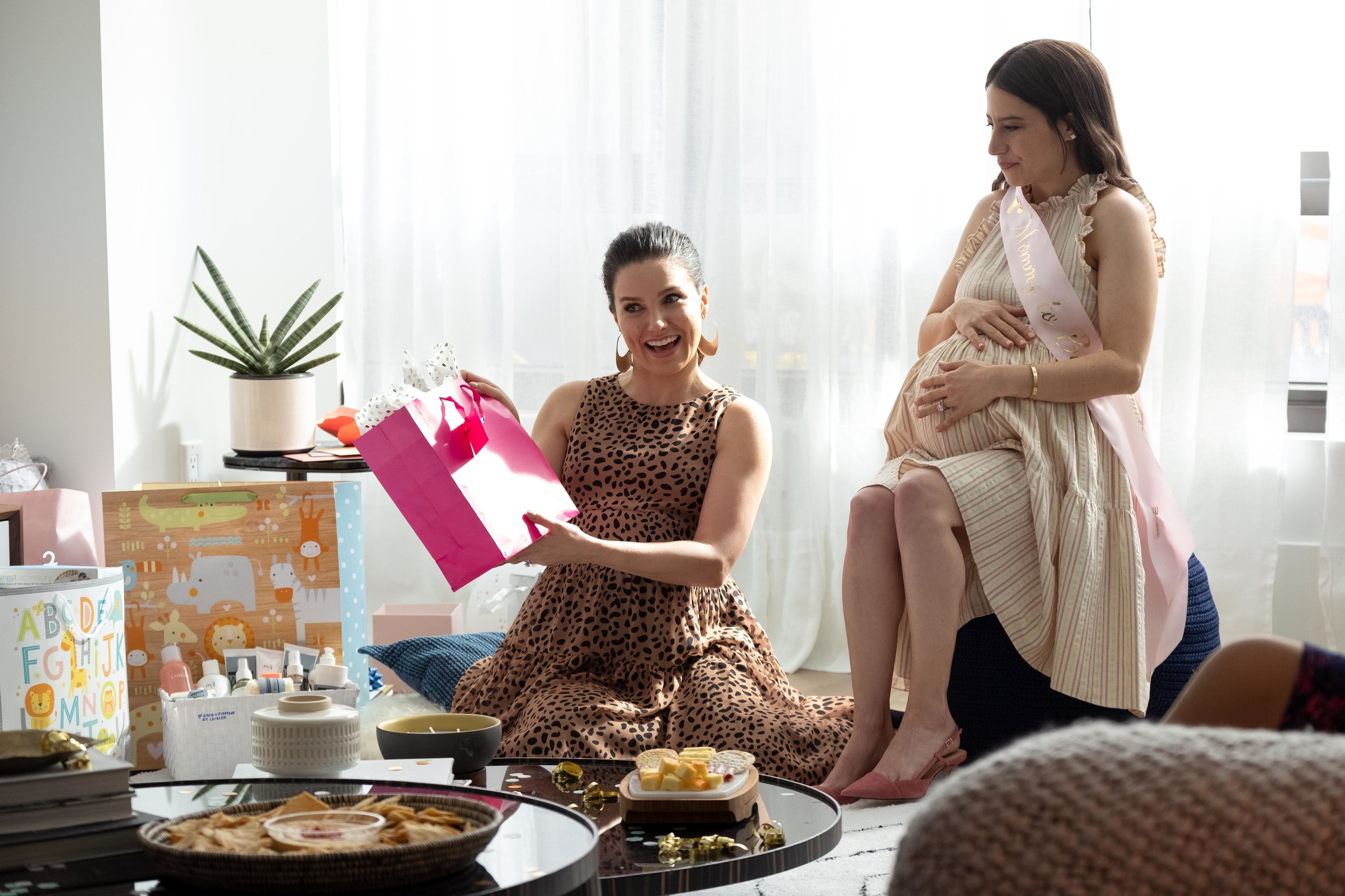 Lucy cradling her baby bump at her baby shower as Sophia&#x27;s character holds up a gift bag