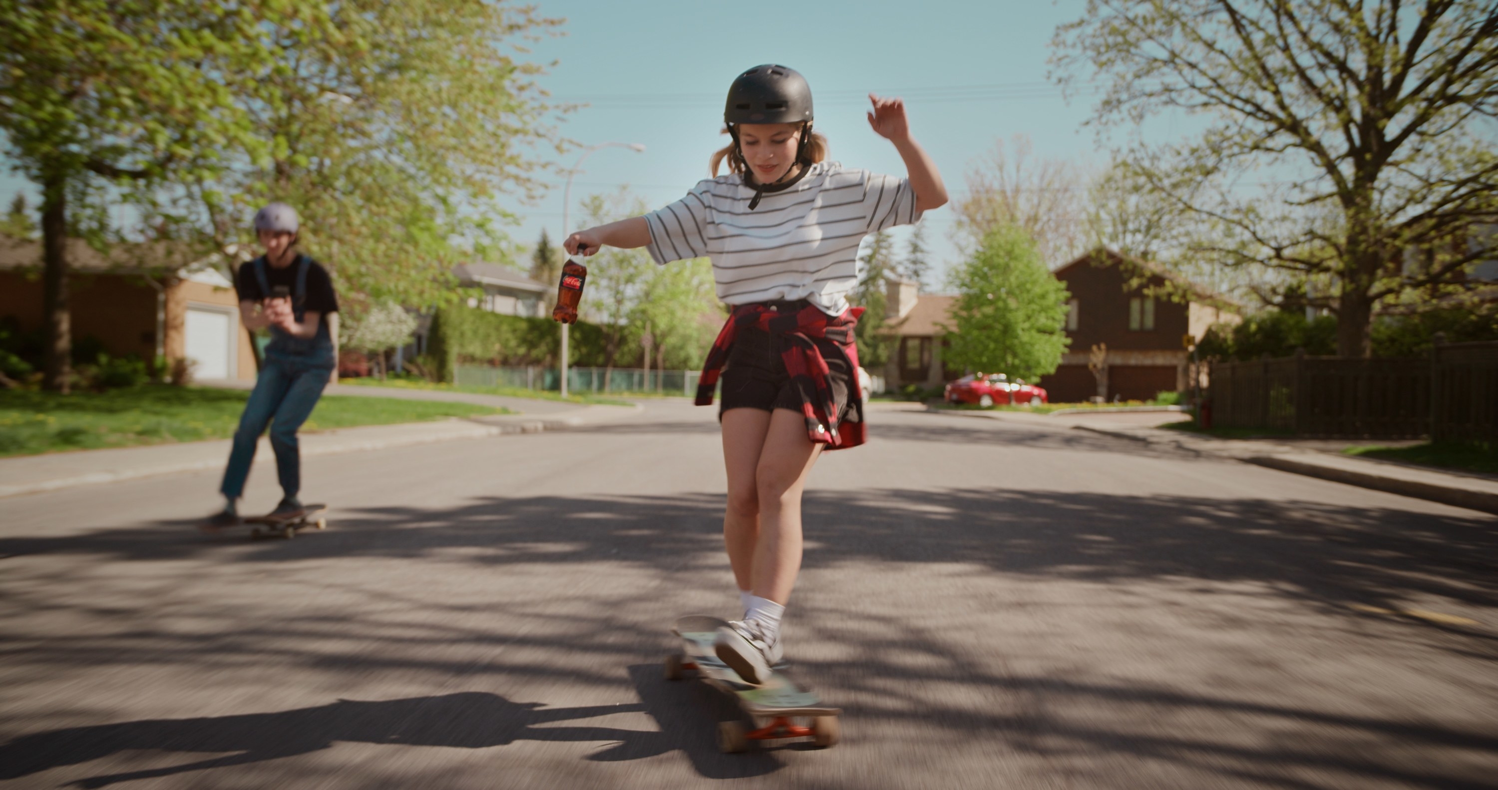 A person skate boarding while holding a Coca Cola mini bottle; their friend is filming on a smartphone