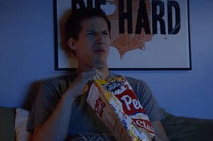 Jake Peralta from Brooklyn99 eating chips. He has a disgusted expression on his face and is reaching inside the bag, right under his face. He is in a dark room, lit by the TV's blue light. He has a "Die Hard" poster behind him and is laying on a bed.
