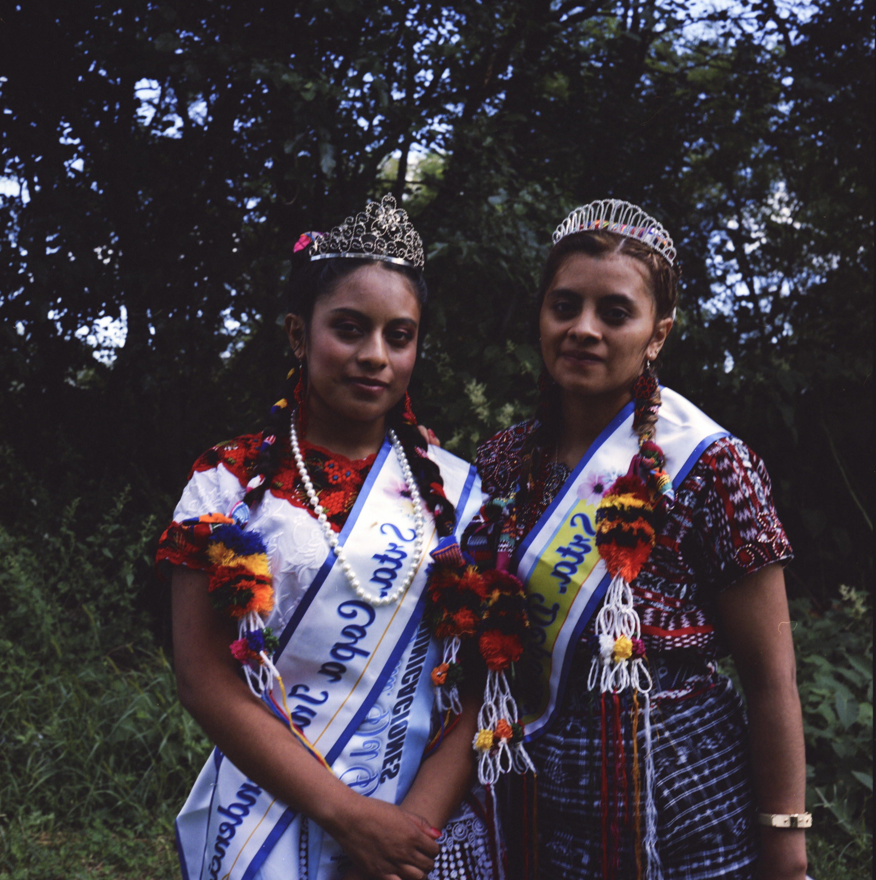 Two women in Indigenous clothing, tiaras, and sashes in front of greenery 