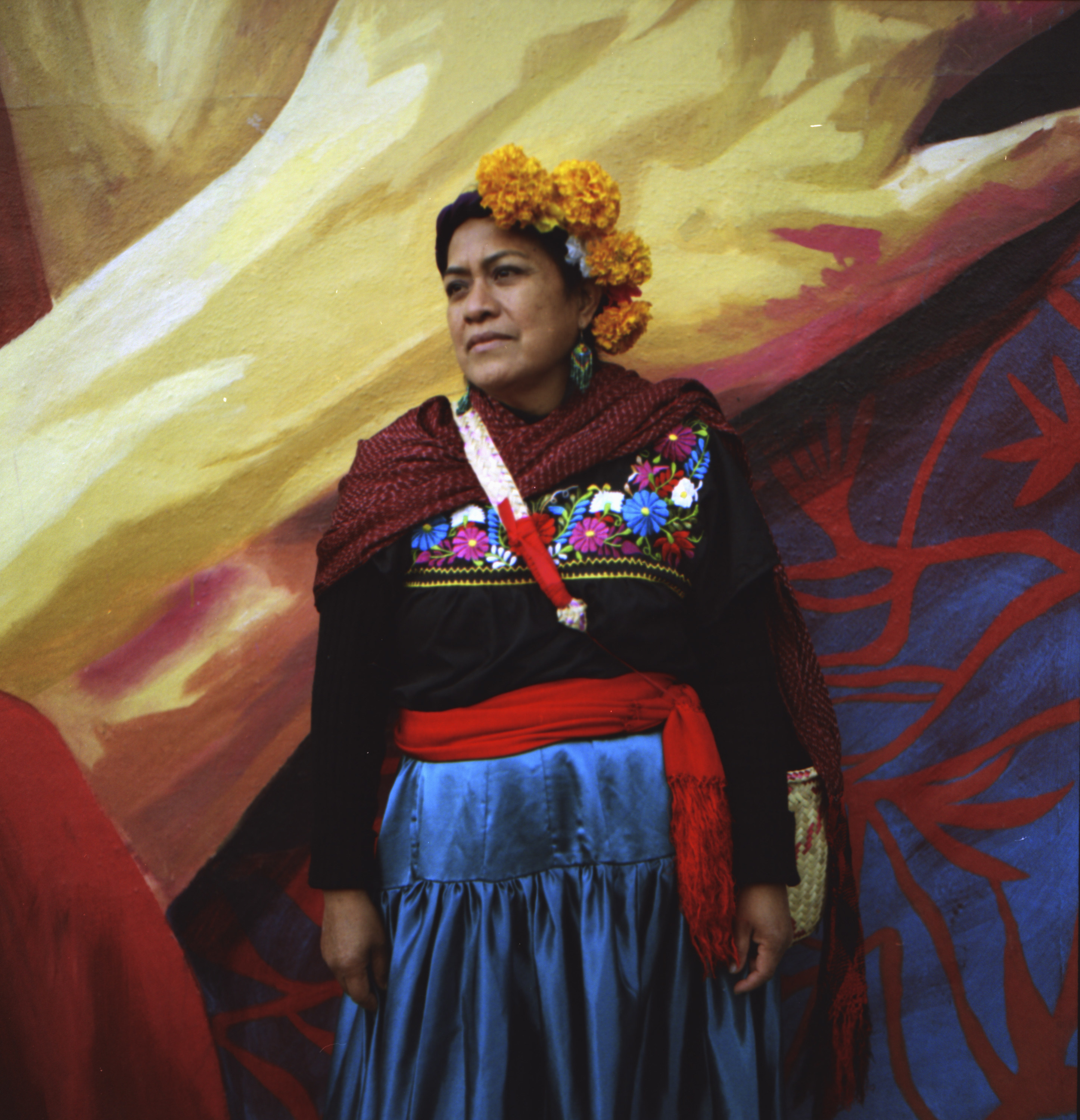 An indigenous Mexican woman with a flower crown standing in front of a mural 