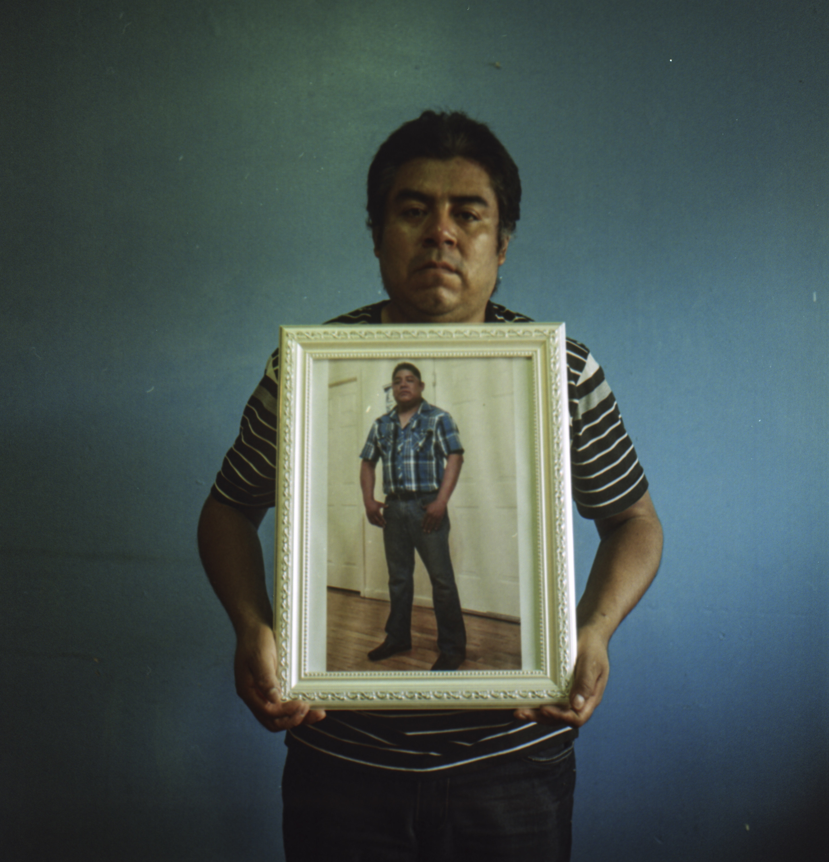 A man holding a framed portrait of his brother on a blue wall background