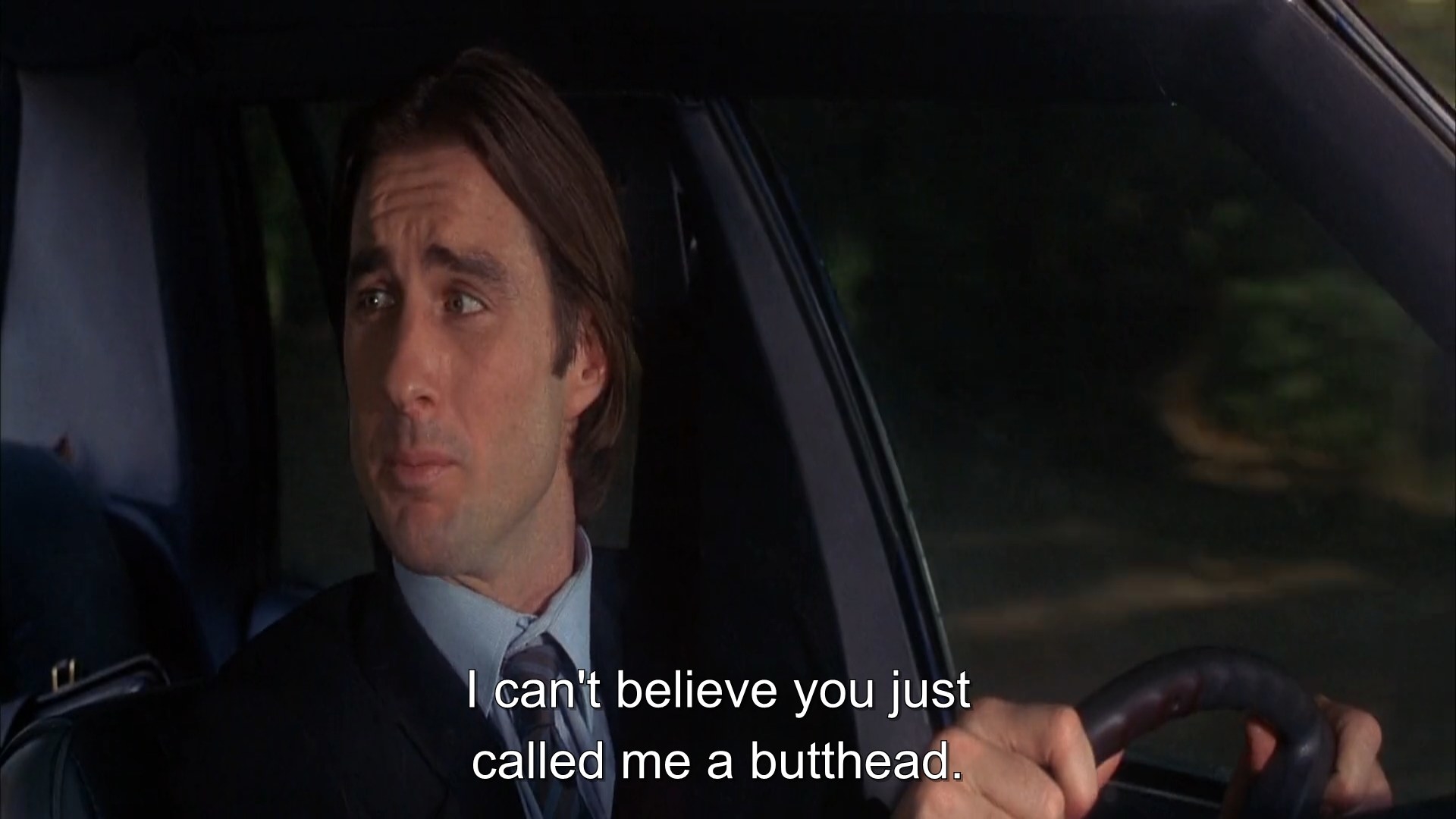 A man in black suit driving a car and looking at his co-passenger and saying, &quot;I can&#x27;t believe you just called me a butthead&quot;