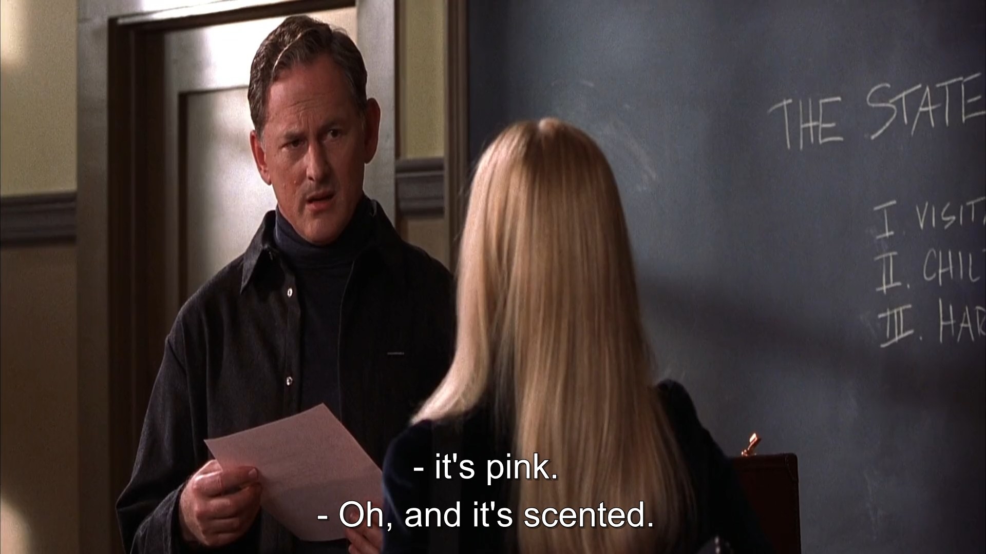 A man in dark turtleneck and jacket holding a pink sheet of paper while he looks on in shock at a blonde girl.