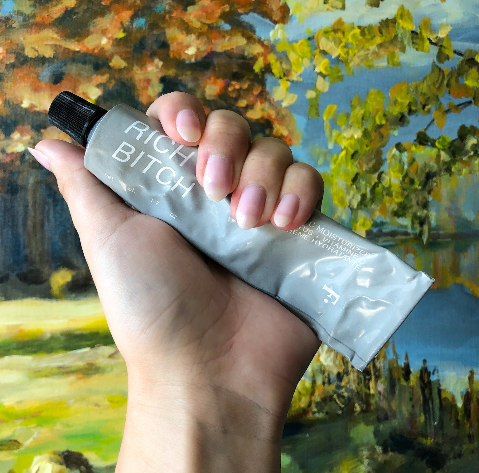 Victoria holding up a tube of the moisturizer against a colourful landscape painting