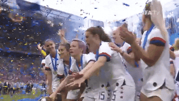 Gold-medal wearing US women&#x27;s soccer team huddled together celebrating on the field amidst confetti and cheers