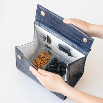 a model opening the navy faux-croc bag showing utensils, a granola bar, and berries inside the lunchbox