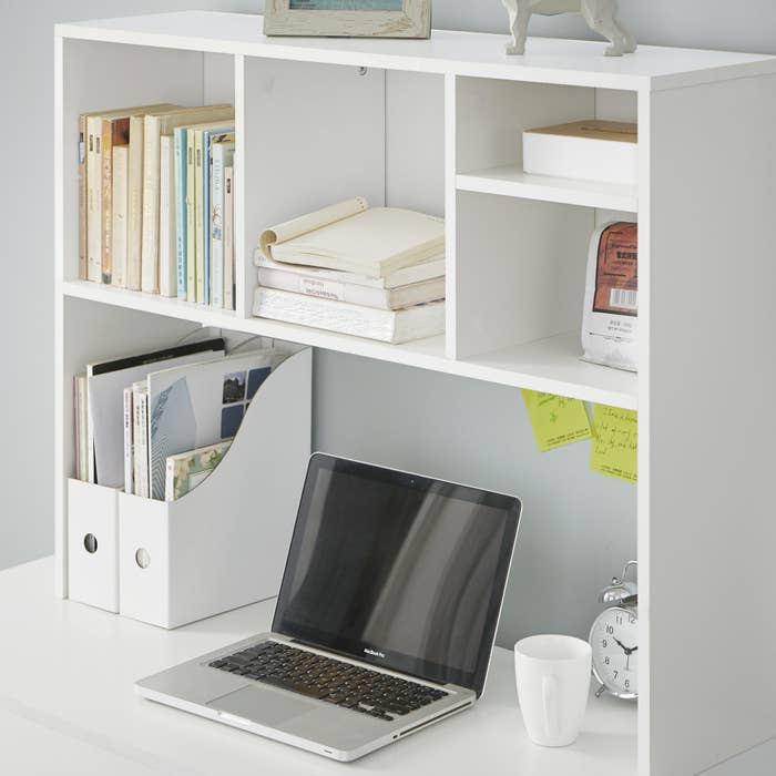White desk with large white desktop riser bookshelf, holding books and other various objects; above a desk with laptop, alarm clock, and other items atop.