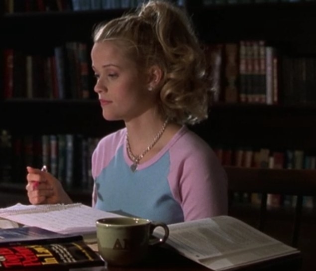 Elle sits at a desk with study materials in front of her, wearing a tee with her hair in a high pony