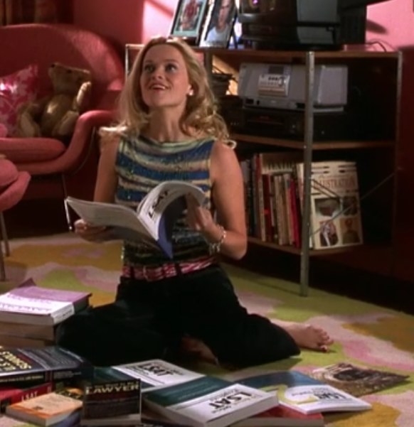 Elle wears a striped top, patterned belt, and dark pants while she sits on the ground surrounding by books