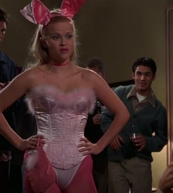 Elle wears a pink Playboy bunny outfit and stands with her hands on her hips