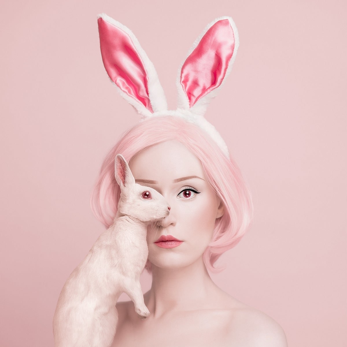 A woman with pink hair wearing bunny ears and holding a rabbit on a pink background 