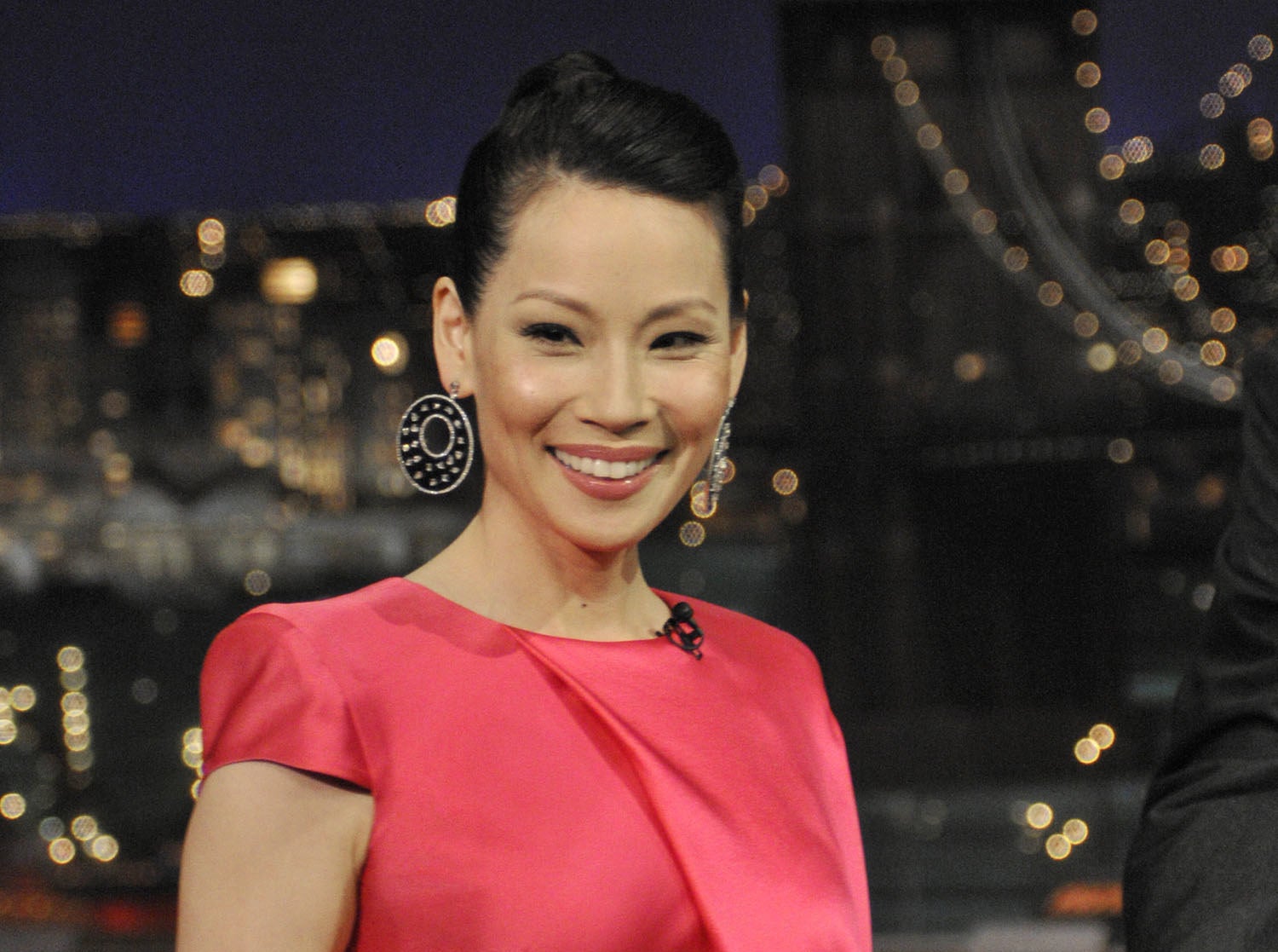 A smiling Lucy Liu wearing large earrings and a short-sleeved dress