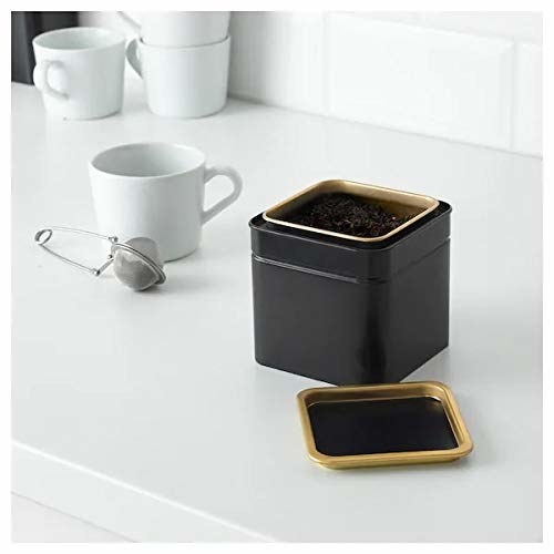 A black and golden tin on a table next to a teacup and a tea infuser ball
