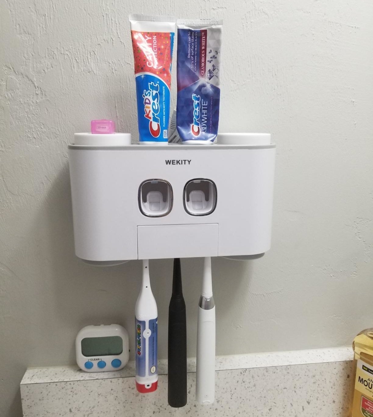 a reviewer photo of the wall-mounted station with three toothbrushes hanging from the bottom and two tubes of toothpaste inserted on the top