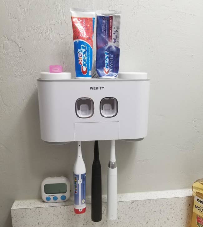 a reviewer's wall-mounted station with three toothbrushes hanging from the bottom and two tubes of toothpaste inserted on the top
