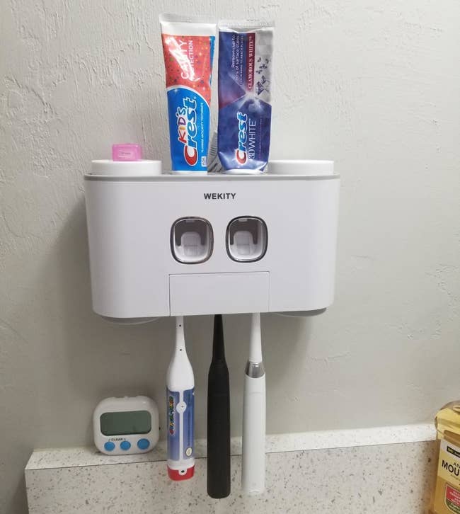 wall-mounted station in a reviewer's bathroom with three toothbrushes hanging from the bottom and two tubes of toothpaste inserted on the top