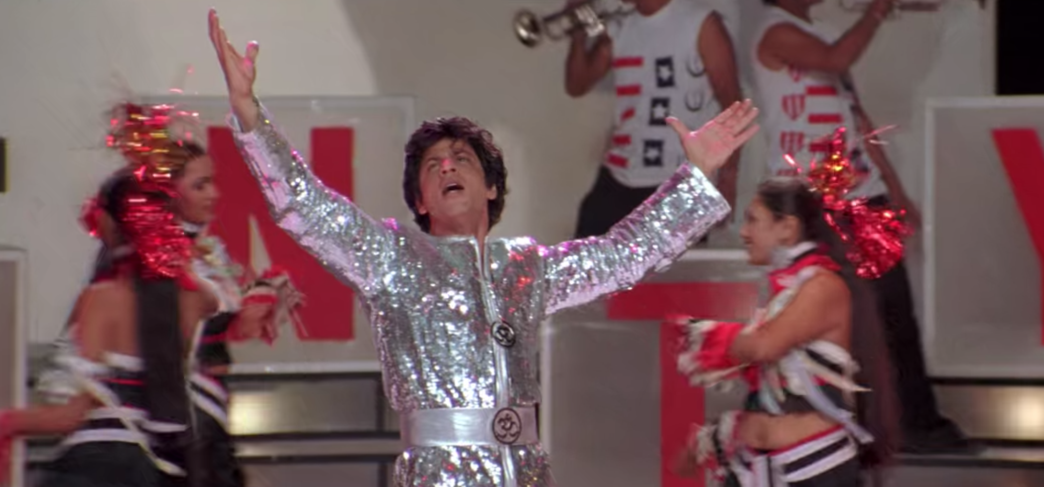 Om wears a sparkly silver jumpsuit as dancers run behind him.
