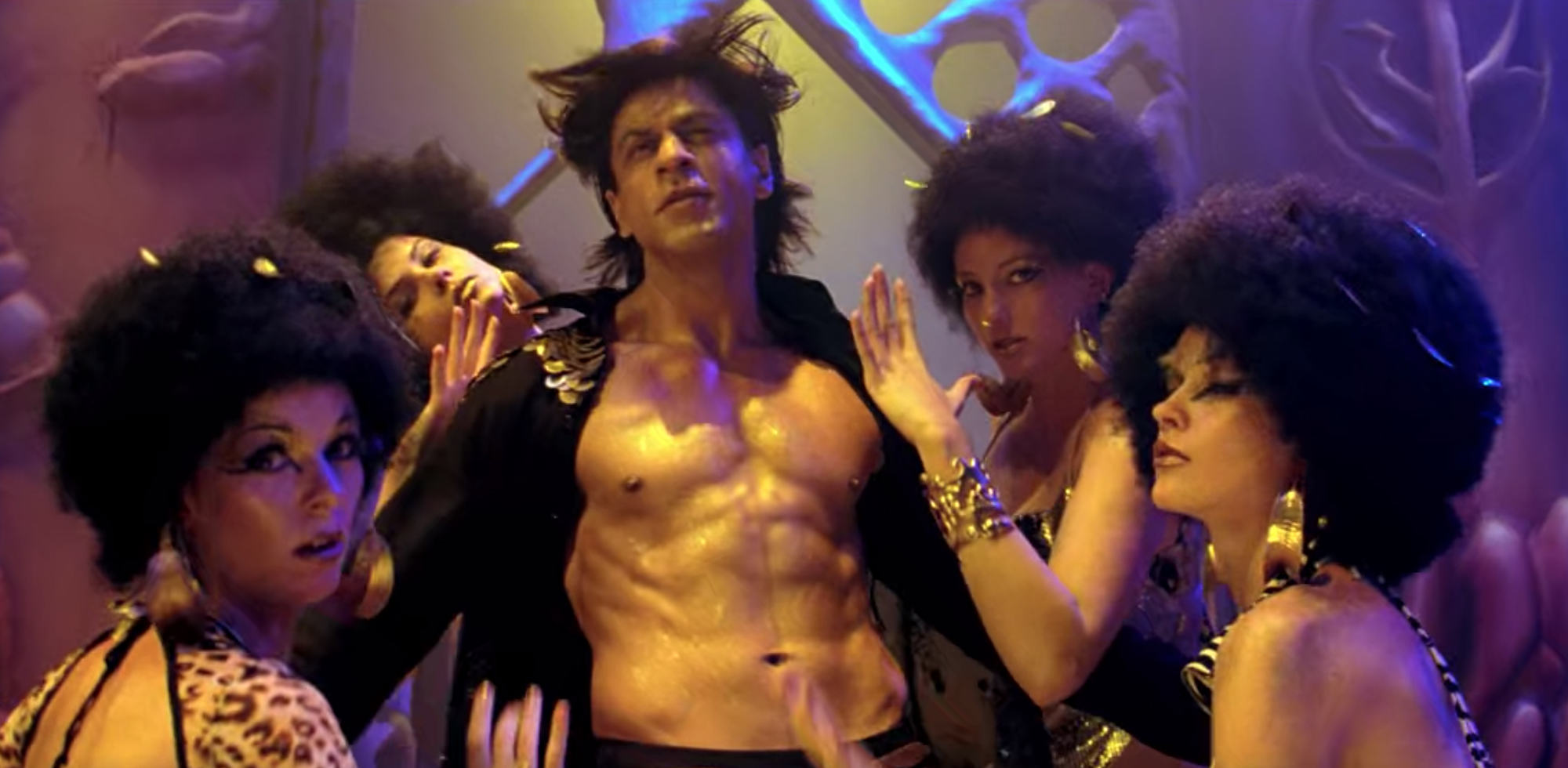Om, shirtless and bathed in golden light, is surrounded by women with afros in cheetah costumes.
