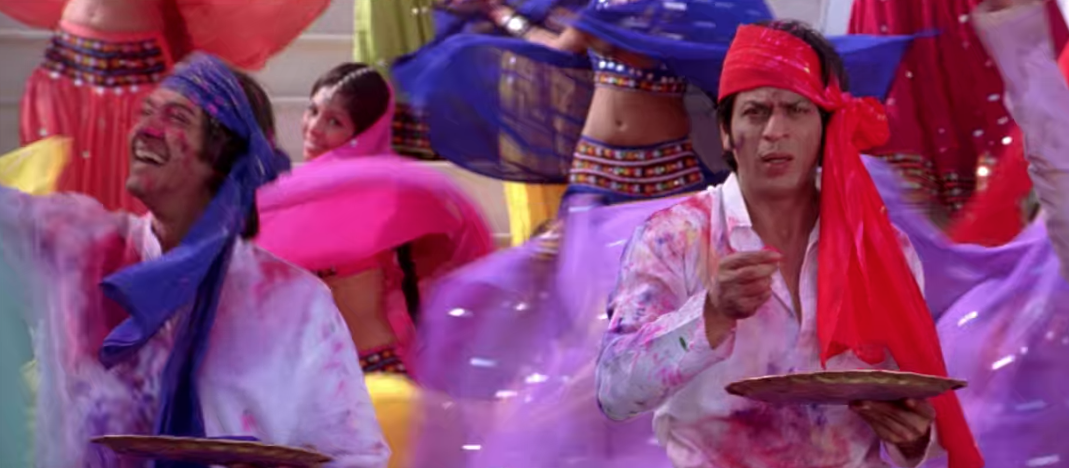 Om, wearing a white shirt and red head scarf, throws colorful dust into the air as women dance behind him.